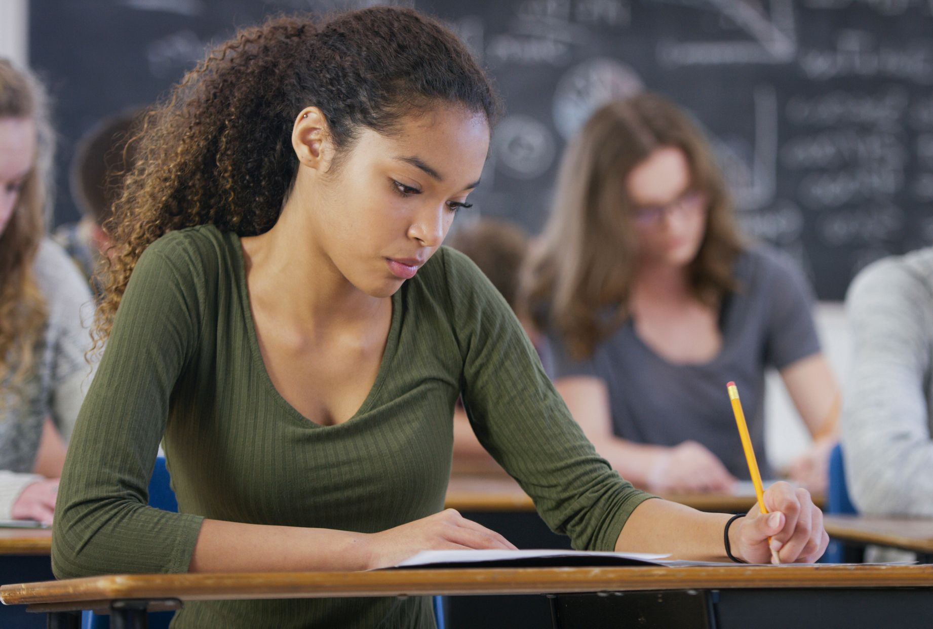 A mixed race teenage girl is taking a high school standardized test in class. She looks down and works on the test.